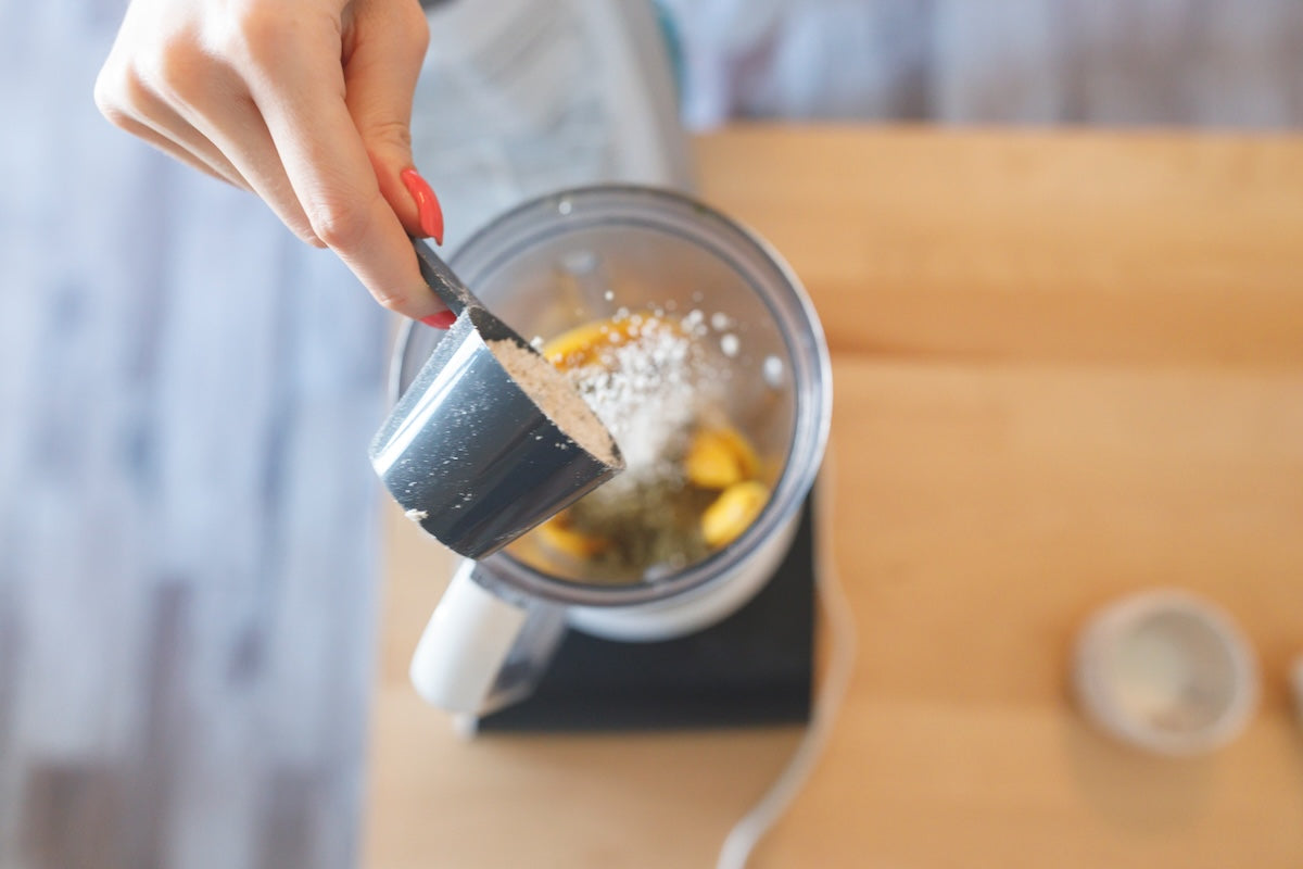 A person’s hand pouring a scoop of white powder into a blender containing yellow ingredients set on a wooden countertop.