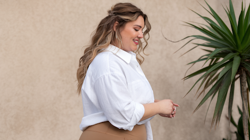 A woman with wavy, light brown hair wearing a white button-up shirt and beige pants, standing in profile against a beige wall with a green plant on the right side.